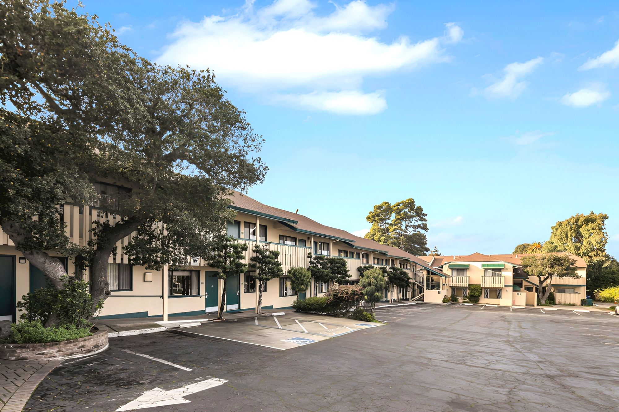 A small motel complex with two-story beige buildings, a paved parking lot, trees, and clear blue sky, under sunny weather.