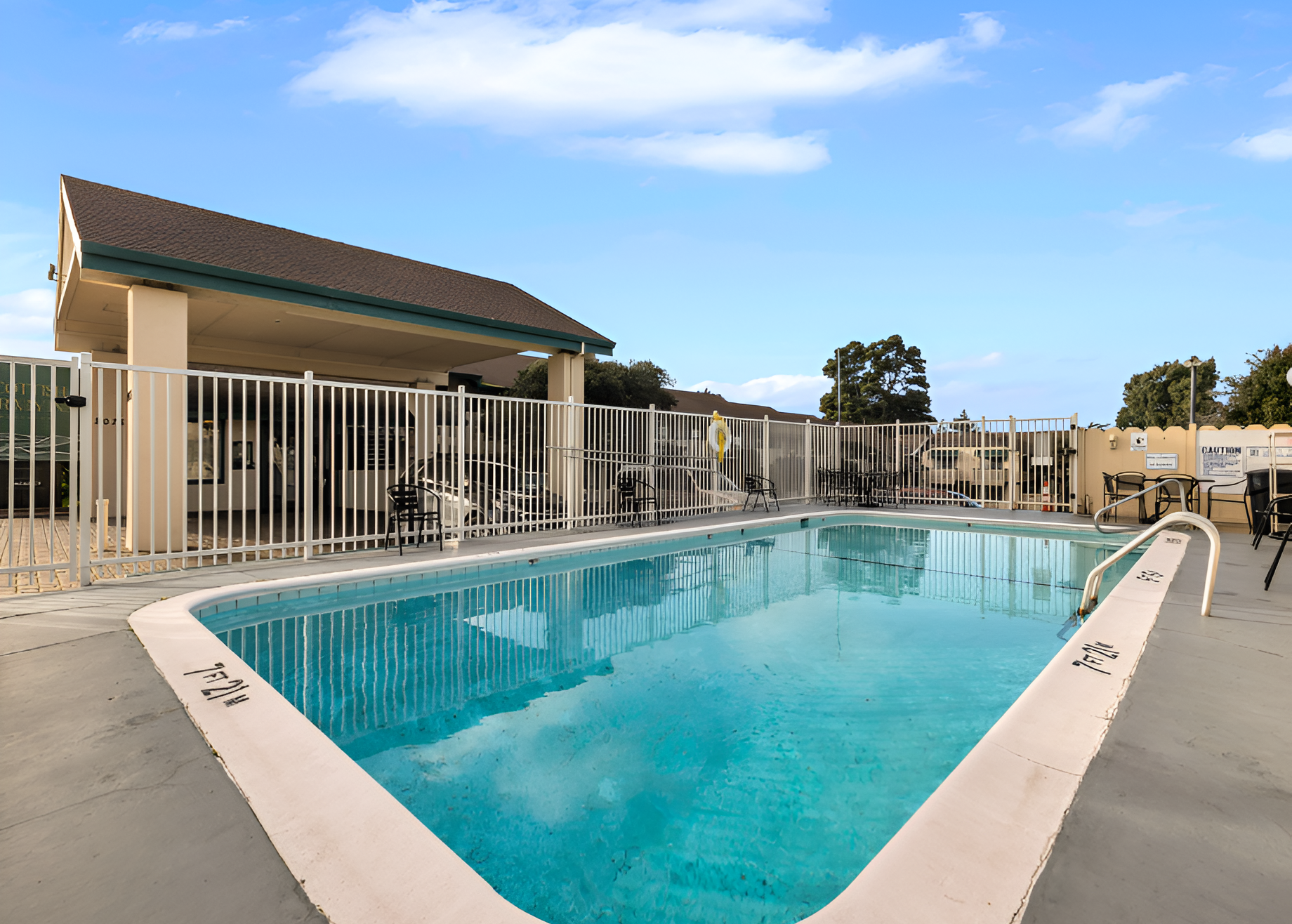 A rectangular outdoor swimming pool with clear blue water, surrounded by a concrete deck and a white fence, in a sunny, apartment-like setting.