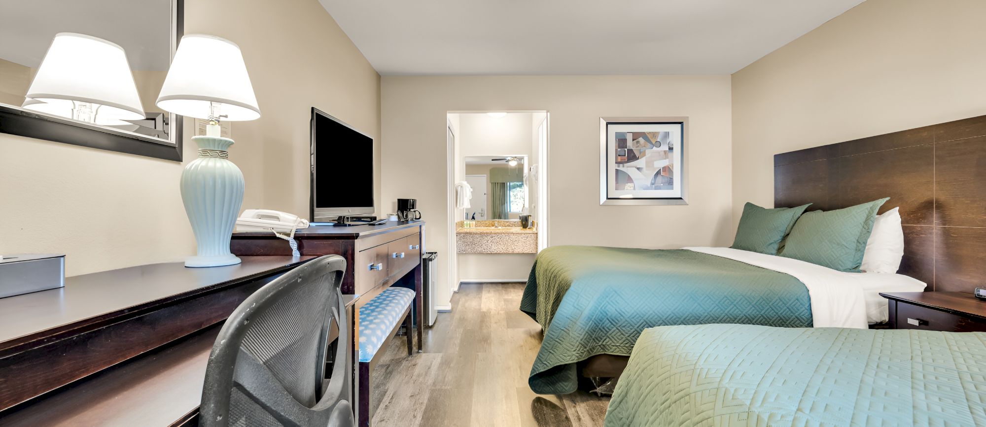 A hotel room with two double beds, green bedspreads, a desk, chair, TV, lamps, and a doorway leading to a bathroom.