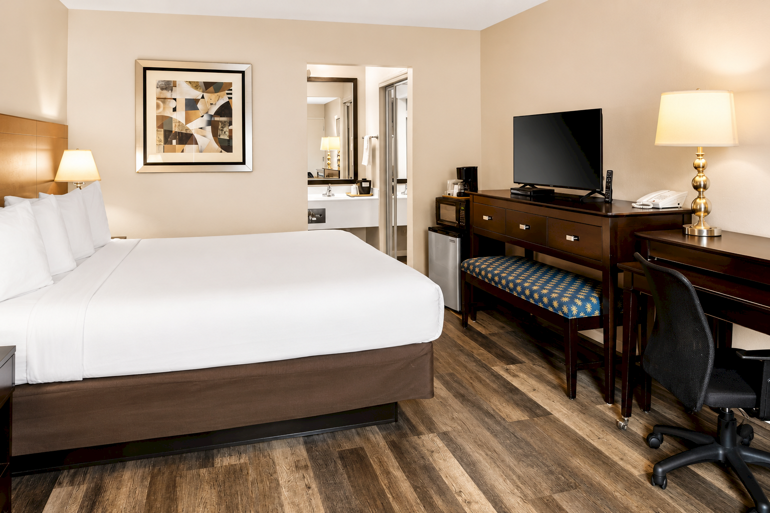 A hotel room with a large bed, wooden furniture, desk and chair, dresser with a TV, bedside lamps, framed artwork, and a doorway to a bathroom; warm lighting.