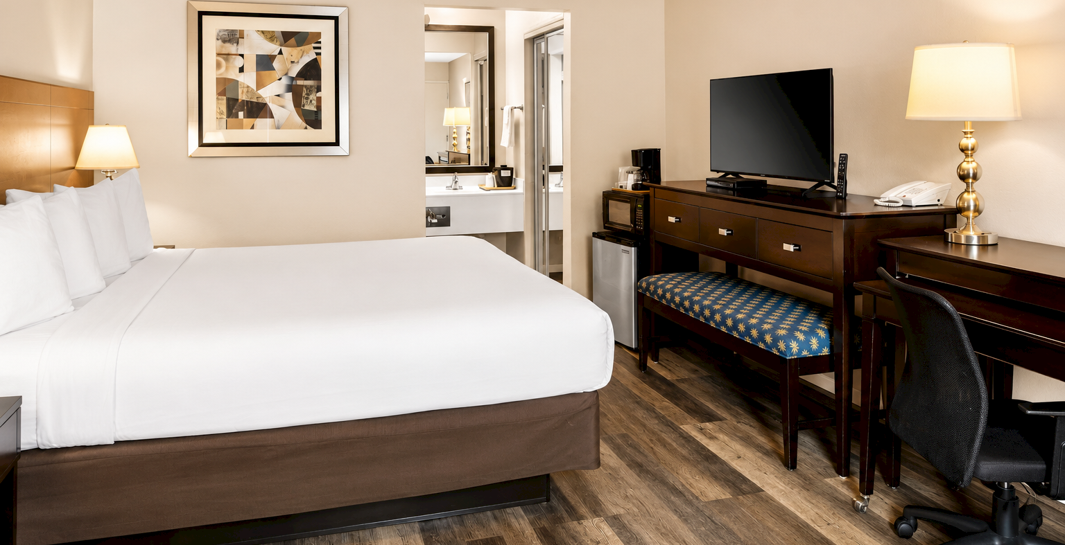 A hotel room with a large bed, wooden furniture, desk and chair, dresser with a TV, bedside lamps, framed artwork, and a doorway to a bathroom; warm lighting.