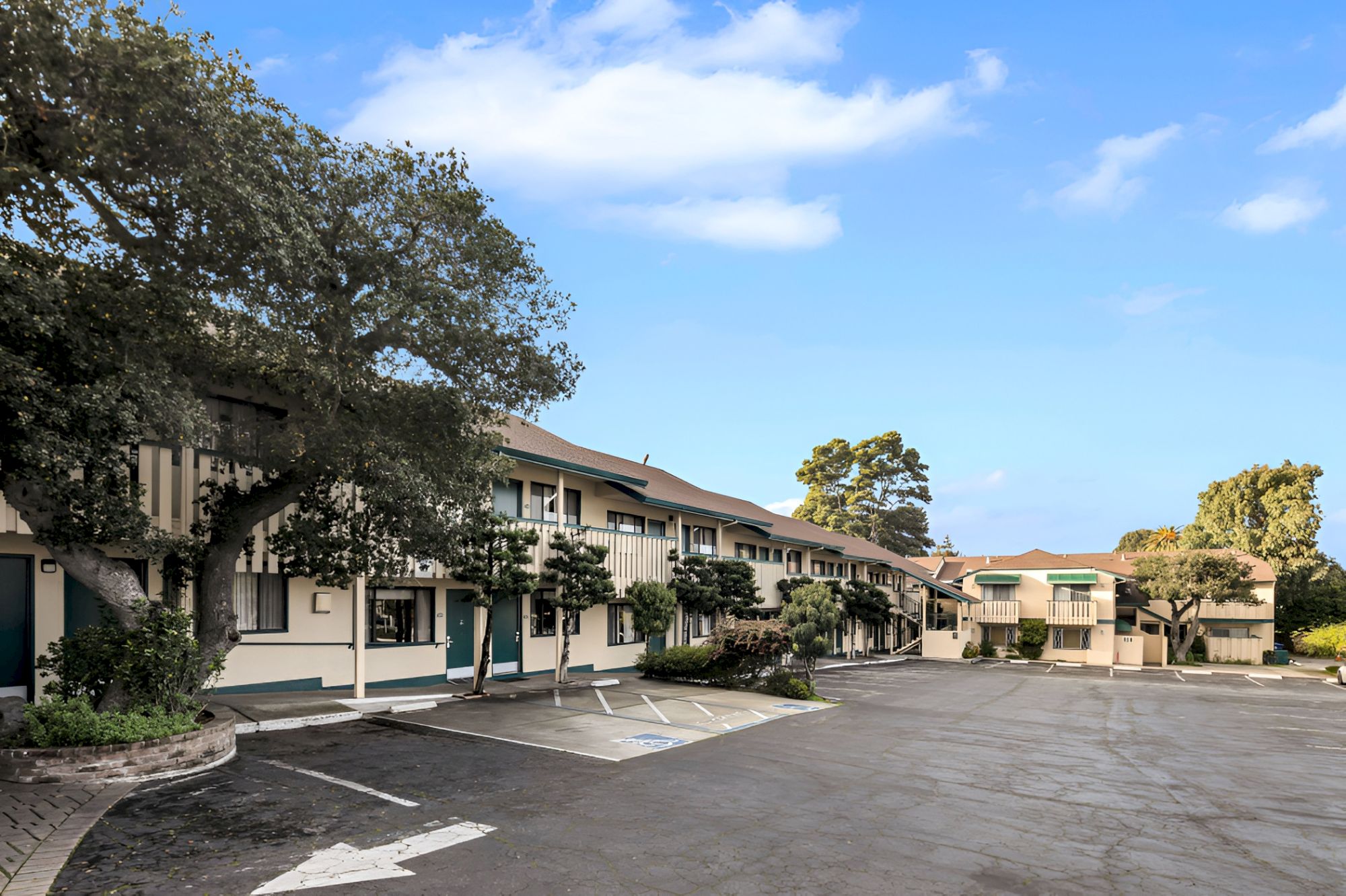 A small, single-story motel or hotel courtyard with a row of cream-colored buildings, parking spaces, trees, and blue sky.