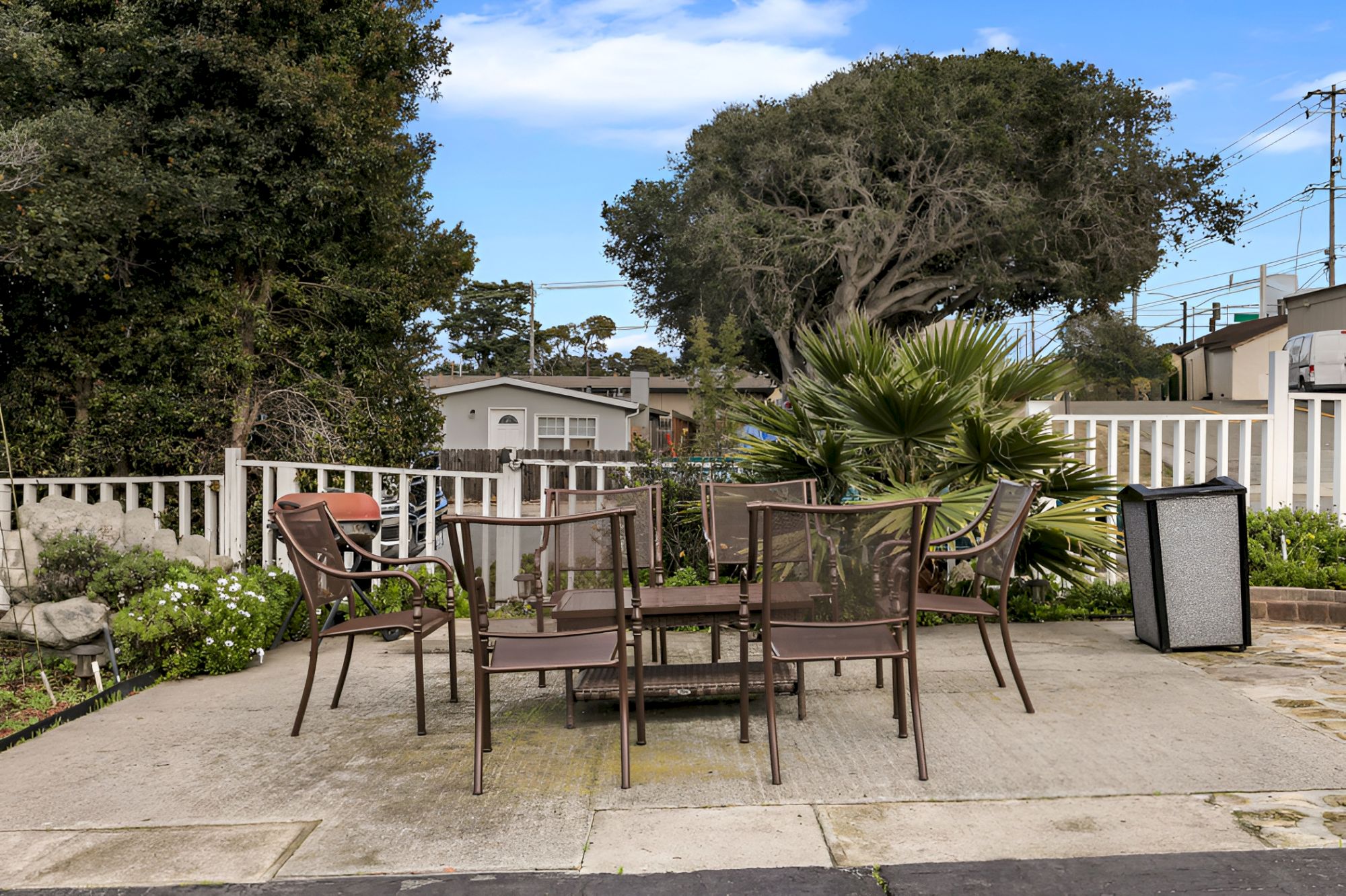 A small outdoor patio with a metal table and six chairs, potted plants, a palm, and a white fence behind on a sunny day.