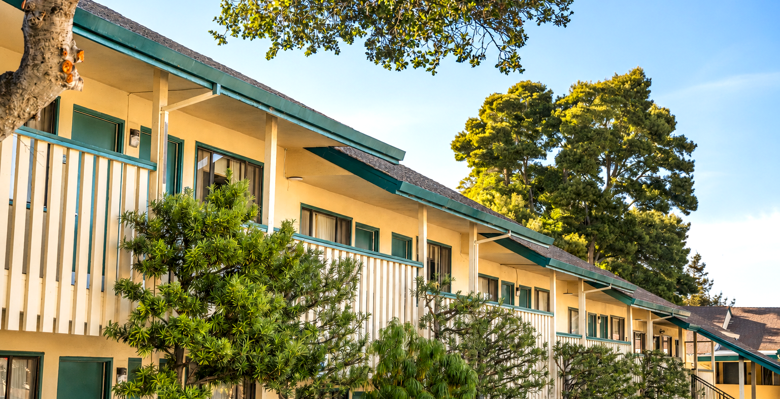 A two-story motel/hotel with yellow exterior walls, green railings, and trees lining the walkway, under a clear sky.