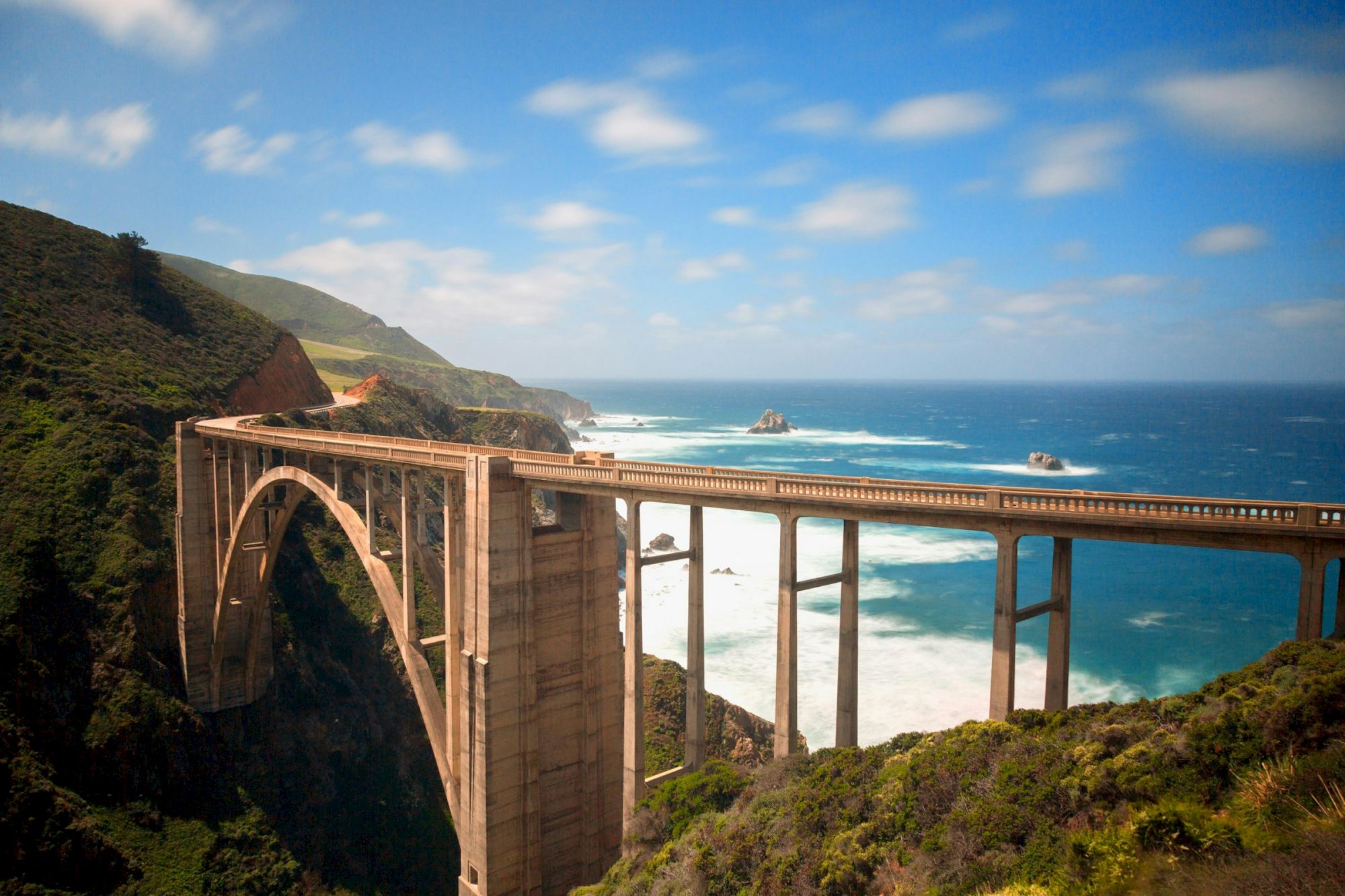 A large stone arch bridge spans a rugged coastline, connecting cliffs over the blue ocean with a bright, partly cloudy sky above.