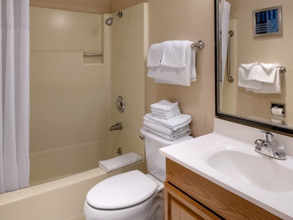 A clean hotel bathroom with a bathtub/shower combo, toilet, a sink with wooden cabinet, and stacked towels; mirror and towels mounted on wall.