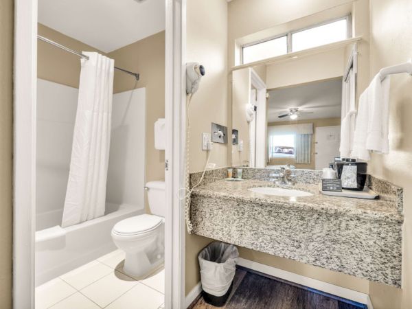 A hotel-style bathroom with a granite double vanity, large mirror, toilet, shower/tub with a white curtain, and towels on a rack, compact and clean.