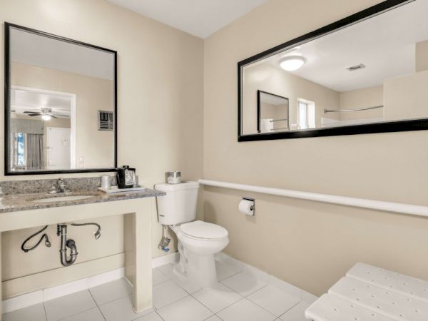 A beige bathroom with a sink and vanity, a toilet, a wall-mounted grab rail, two mirrors, and a white tiled floor.