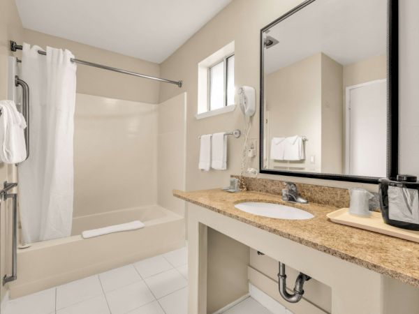 A clean, beige bathroom with a tub/shower, white tiles, a granite counter, twin sinks, and a large mirror; towels and essentials arranged neatly.