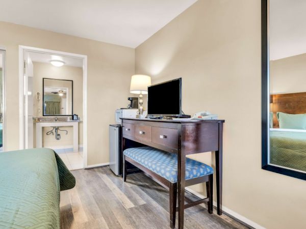A hotel room with a bed in the foreground, a desk with a flat-screen TV, a bench chair, a large wall mirror, and a doorway to a bathroom.