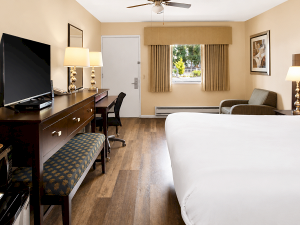 A hotel room with a large bed, desk and chair, flat-screen TV, dresser, two lamps, a bench, and a window with beige curtains.