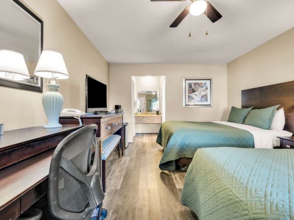 A hotel room with two double beds, a desk and chair, a wall-mounted TV, a dresser, and a ceiling fan; carpeted floors and warm lighting.