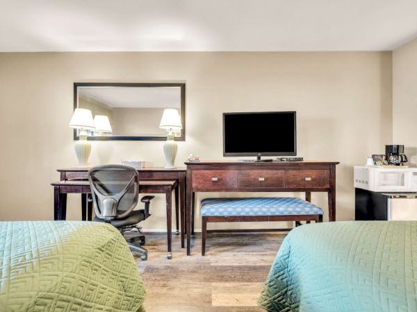 A hotel room with two green-banded beds, a desk and chair, a wall mirror, a dresser with a small TV, and a coffee/tea station.