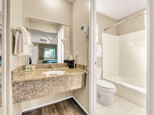 A hotel-style bathroom with a granite counter, double sink, mirror, towels, toilet, and a tub/shower combo.