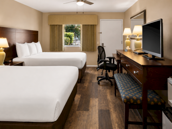 Two beds in a hotel room with a desk, chair, lamps, a window with blinds, and a flat-screen TV on a long dresser.