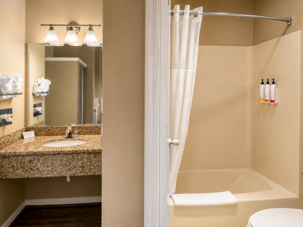 A hotel bathroom with a granite vanity, large mirror, and two lights; there&rsquo;s a separate shower/tub with a white curtain, towels, and bath products.