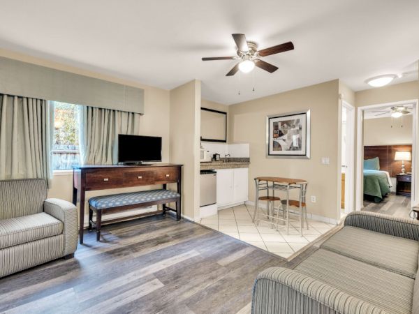 A cozy hotel suite living area with a TV on a dresser, sofa and chair, a small kitchenette with bar seating, and a doorway to a bedroom, all in neutral tones.