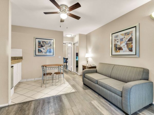 A cozy living area with a gray sofa, wall art, ceiling fan, and a small dining nook near a kitchenette; hallway leads to more rooms.