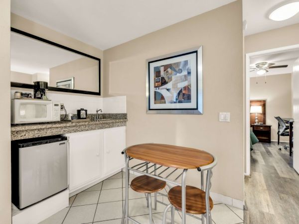 A compact kitchenette with white cabinets, granite countertop, mini-fridge, microwave, and a small round table with two stools near a beige wall.