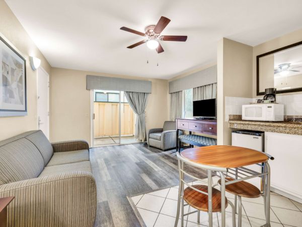 A small living area with a beige sofa, a wall art frame, a ceiling fan, a kitchenette with a granite counter, a round wooden table with two chairs, and sliding glass doors leading to an outside patio.