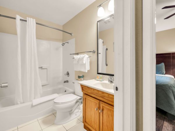 A compact bathroom with a bathtub/shower combo, toilet, and wooden vanity with a single sink; glimpse of a bedroom beyond.
