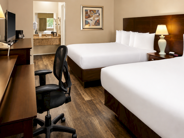 A hotel room with two queen beds, a desk and chair, a lamp, and a mirror reflecting a seating area. End.