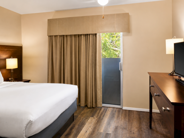 A tidy hotel room with a king bed, nightstand lamps, a desk with a TV, and beige walls; a windowed door leads to a balcony.