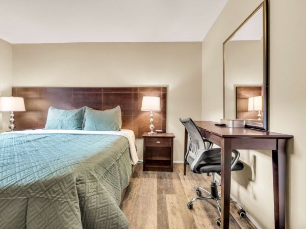 A hotel room with a king bed, teal quilt, two matching pillows, nightstands with lamps, and a desk with chair and a large wall mirror.