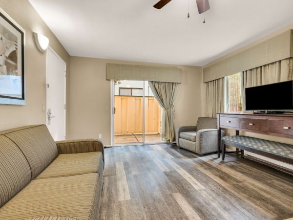 A cozy living room with a tan sofa, striped armchairs, wood floor, and a long wooden console with a TV; sliding door to an outdoor area.
