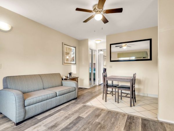 Cozy living area with a gray sofa, small dining table, ceiling fan, and a doorway leading to another room&mdash;bright, simple, and inviting.