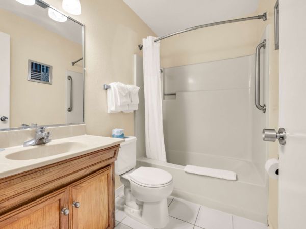 A clean bathroom with a wooden vanity, mirror, toilet, and a white-tiled shower with a curved rod and curtain, plus a towel rack and a small trash can.