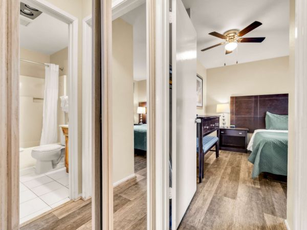 A hotel hallway with a view into a bedroom and an adjacent bathroom, featuring a ceiling fan, wooden floors, and teal beds, across a mirrored hallway.
