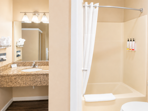 A hotel bathroom with a tan granite vanity, mirror and light fixture, plus a separate beige-tiled shower/bathtub area with a white curtain and towels.