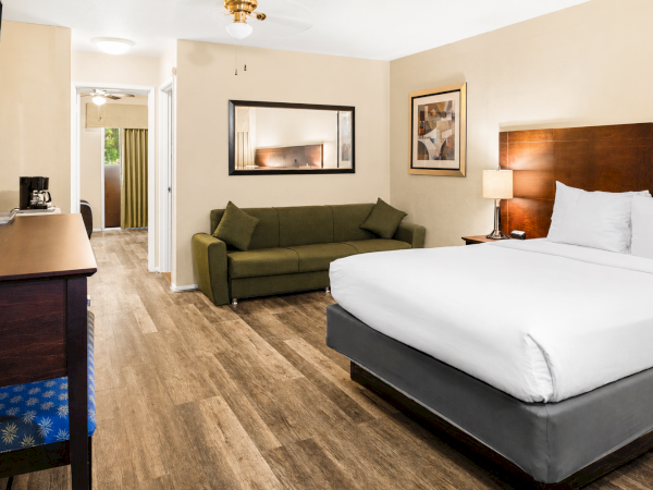 Hotel room with a king bed, gray base, green sofa, wooden floors, desk, flat-screen TV, framed art, and a doorway to a hallway.