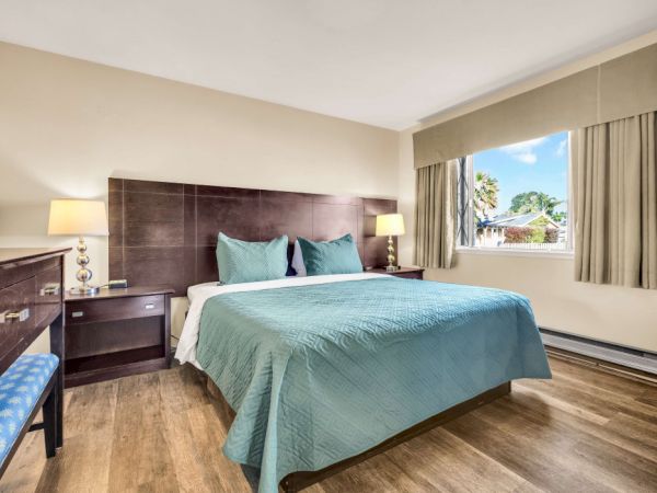 A cozy hotel room with a king bed, teal bedspread, matching pillows, wooden nightstands, two lamps, a window with curtains, and wood-look flooring.