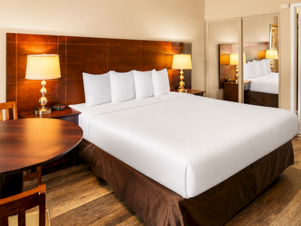 A clean hotel room with a king bed, white linens, matching pillows, wooden headboard, nightstands with lamps, a round table, and mirrored wardrobe.
