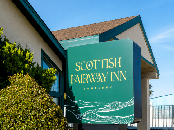 A teal Scottish Fairyway Inn sign on a small building, with plants and a brick border along a sunny sidewalk.