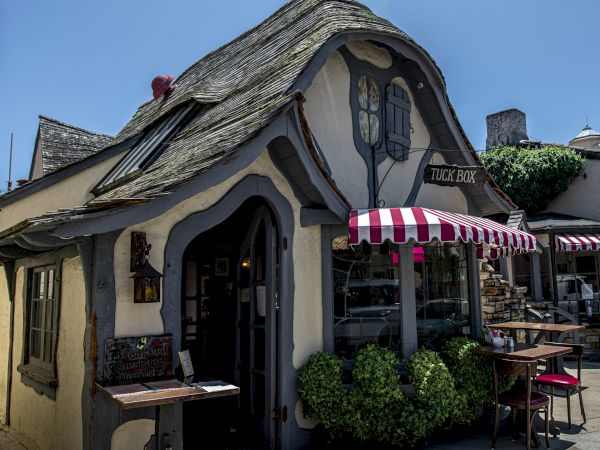 A whimsical, cottage-style cafe with curved, fairy-tale architecture, outdoor seating, striped awning, and stone path, inviting pastry vibes.