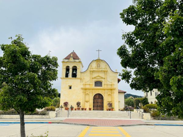 A yellow church with a bell tower, set in a plaza framed by green trees and a clear sky, welcoming visitors with a brown arched door.