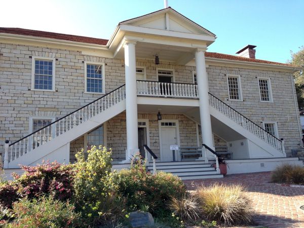 A two-story beige brick building with white columns, a central entry with stairs and a small porch, and well-kept landscaping in front.