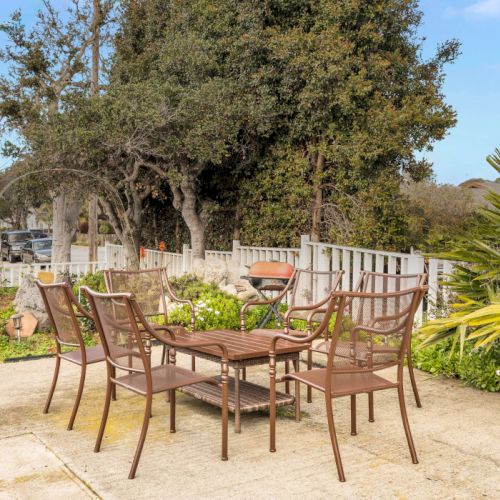 A patio with a round table and six metal chairs, potted plants, a grill in the background, and a garden with trees under a sunny sky.