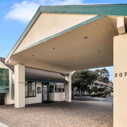 A hotel entrance with a large, sloped canopy, beige walls, and a green sign reading &ldquo;Scottish Inn&rdquo; beside the driveway. The address 2075 is visible on a pillar.