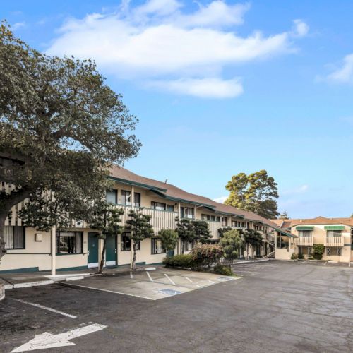 Stray row of beige motel-style buildings with a parking lot, trees, and a bright blue sky scattered with a few clouds, calm and suburban.
