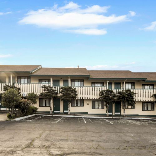 A two-story motel with exterior doors to rooms, a wide parking lot, small trees, and a bright blue sky above the beige building. End.