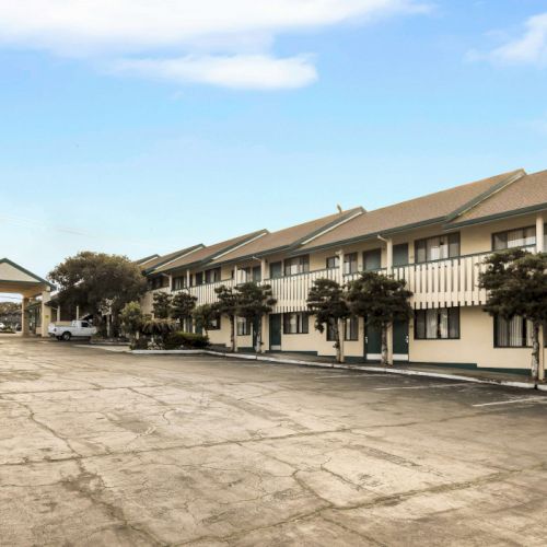 A row of two-story motel/inn buildings with a large empty parking lot in front, under a partly cloudy sky.
