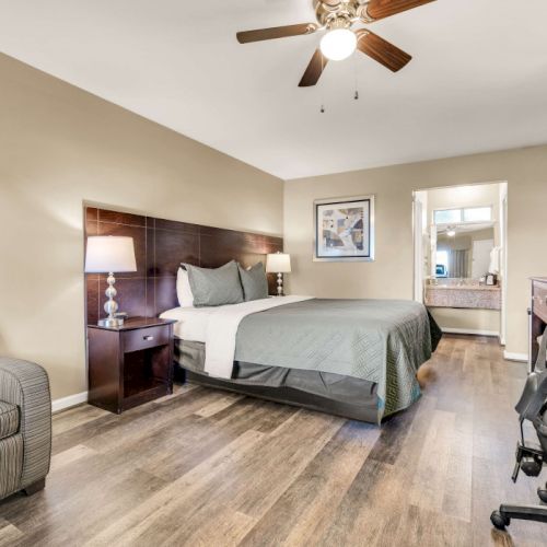 A cozy hotel-style bedroom with a king bed, nightstands, armchair, desk, and a view toward an en suite bathroom, all in warm tones.