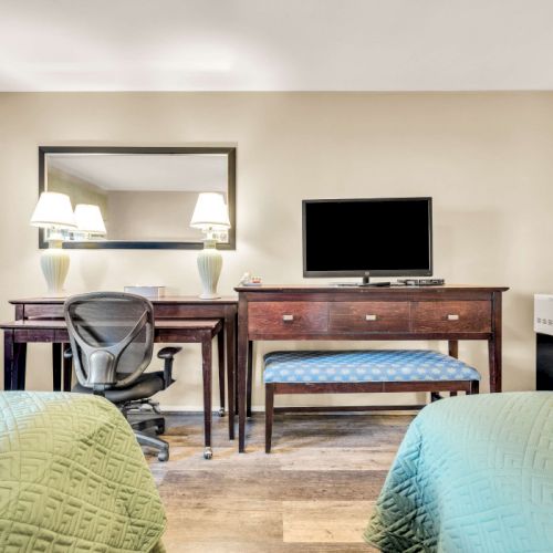 A hotel room with two green bedspreads, a wooden desk and chair, a wall mirror, a dresser with a TV, and a small coffee/kitchen area on the right.