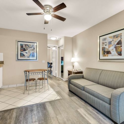 A compact living area with a gray sofa, wall art, ceiling fan, and a small dining table near a tiled kitchenette&mdash;bright, cozy, and modern.