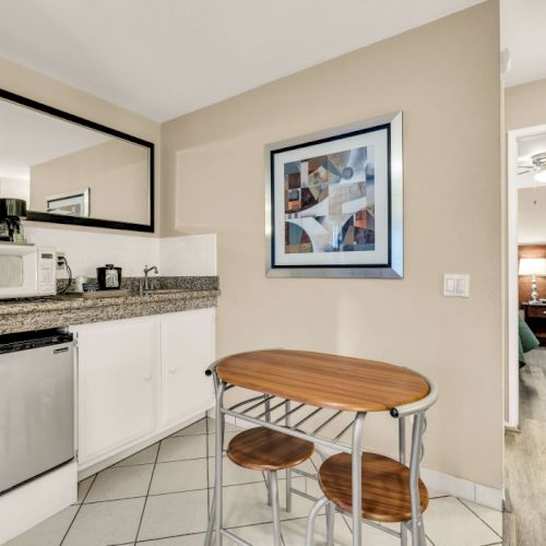 A small kitchenette with white cabinets, a granite counter, mini fridge, microwave, and a round wooden table with two stools. Also a framed art piece on the wall and a doorway to another room.