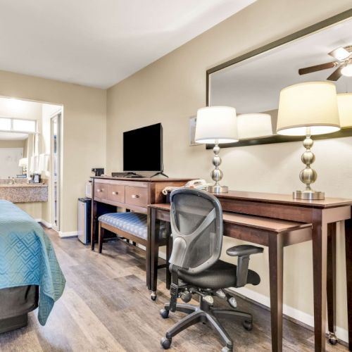 A tidy hotel room workspace beside a bed: desk with chair, dual lamps, wall mirror, TV on a dresser, open doorway to a kitchenette area, warm lighting.